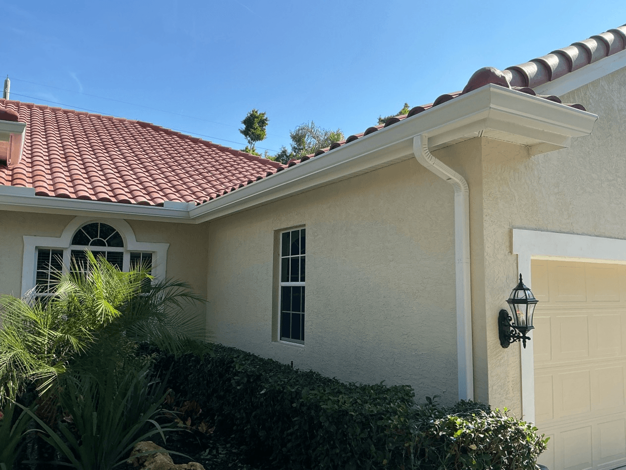 An off-white stucco house with red roof tiles and new white gutters and downspout.